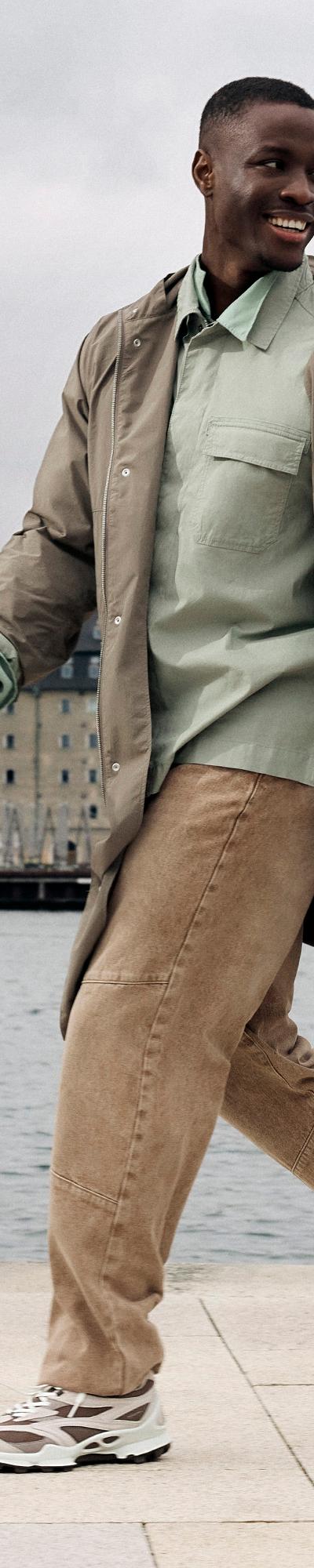 A man walking along a Copenhagen waterfront in a taupe trench coat, sage overshirt and chunky white Ecco sneakers