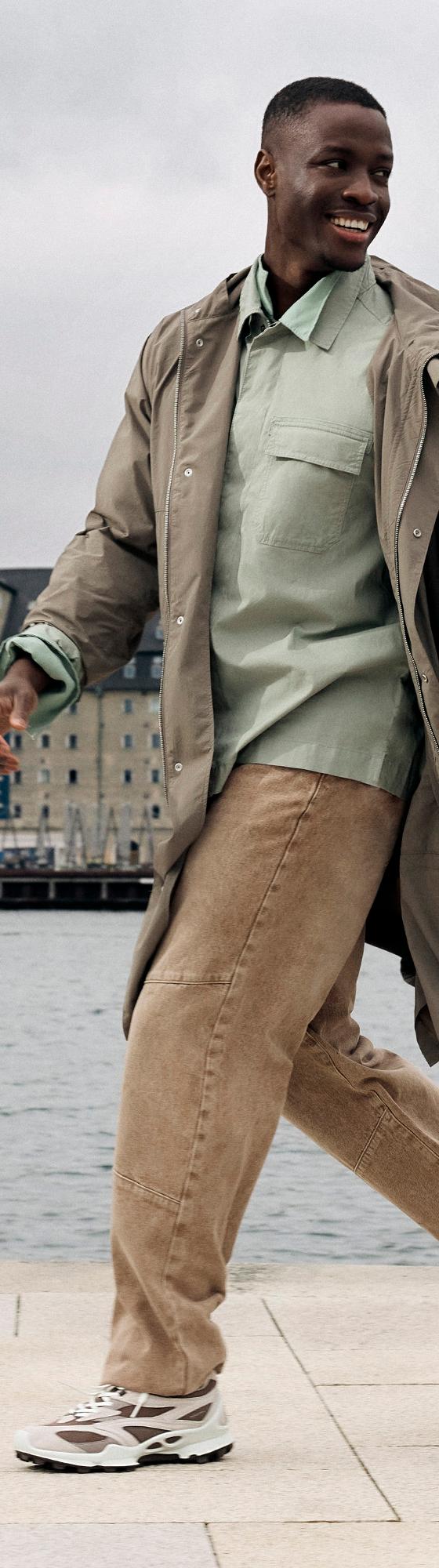 A man walking along a Copenhagen waterfront in a taupe trench coat, sage overshirt and chunky white Ecco sneakers