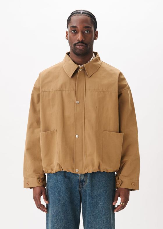 A man wearing a tan Estudio Niksen workwear jacket with blue jeans