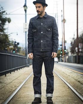 A man wearing an indigo F.O.B FACTORY denim jacket and cuffed selvedge jeans, standing on tram tracks