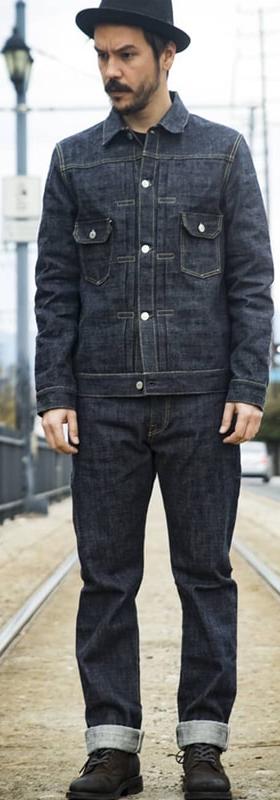 A man wearing an indigo F.O.B FACTORY denim jacket and cuffed selvedge jeans, standing on tram tracks