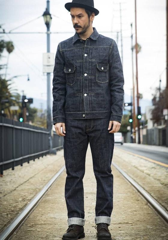 A man wearing an indigo F.O.B FACTORY denim jacket and cuffed selvedge jeans, standing on tram tracks