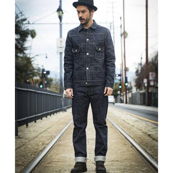 A man wearing an indigo F.O.B FACTORY denim jacket and cuffed selvedge jeans, standing on tram tracks