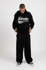 A man wearing a black Federation hoodie with a white script logo, wide black pleated trousers and black canvas sneakers