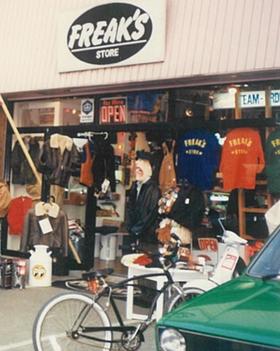 Archive shopfront: FREAK'S STORE sign, racks of jackets and tees spilling onto the street, a man in the doorway, bicycle in front