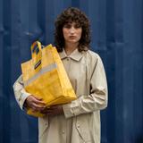 A woman in a beige trench coat carrying a yellow FREITAG tarpaulin tote bag against a blue shipping container