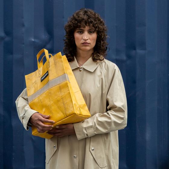 A woman in a beige trench coat carrying a yellow FREITAG tarpaulin tote bag against a blue shipping container