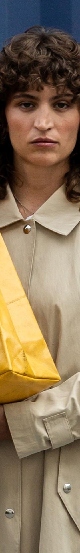 A woman in a beige trench coat carrying a yellow FREITAG tarpaulin tote bag against a blue shipping container