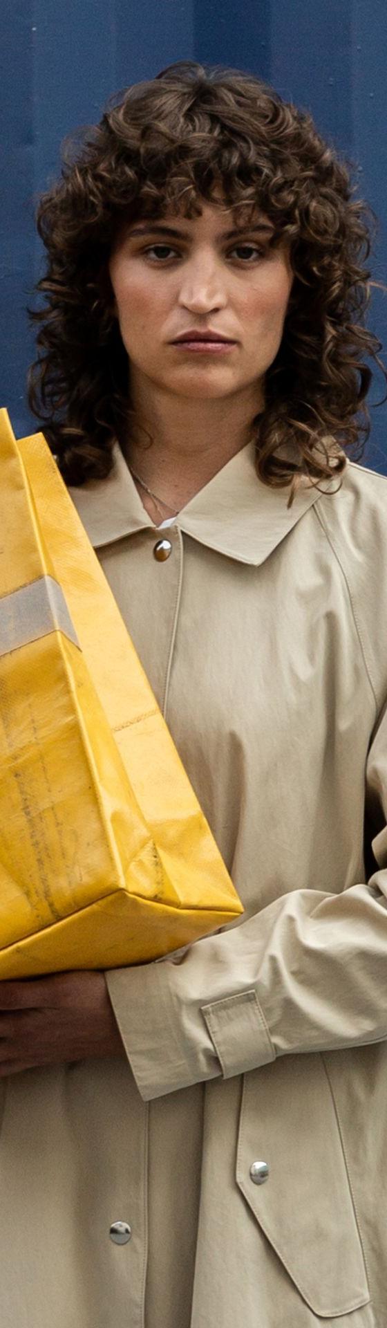 A woman in a beige trench coat carrying a yellow FREITAG tarpaulin tote bag against a blue shipping container