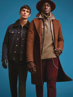 Two men wearing tailored outerwear, one in a camel wool overcoat with a wide-brim hat and the other in a dark denim jacket