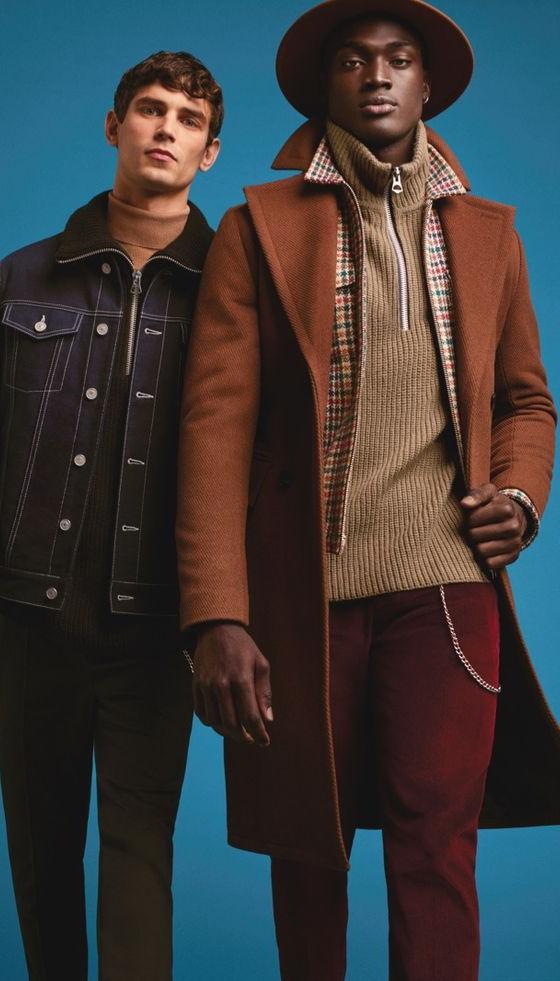 Two men wearing tailored outerwear, one in a camel wool overcoat with a wide-brim hat and the other in a dark denim jacket