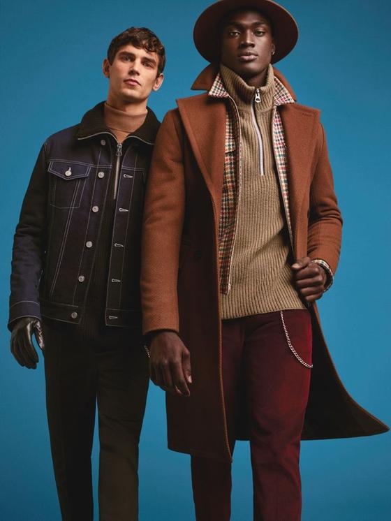Two men wearing tailored outerwear, one in a camel wool overcoat with a wide-brim hat and the other in a dark denim jacket