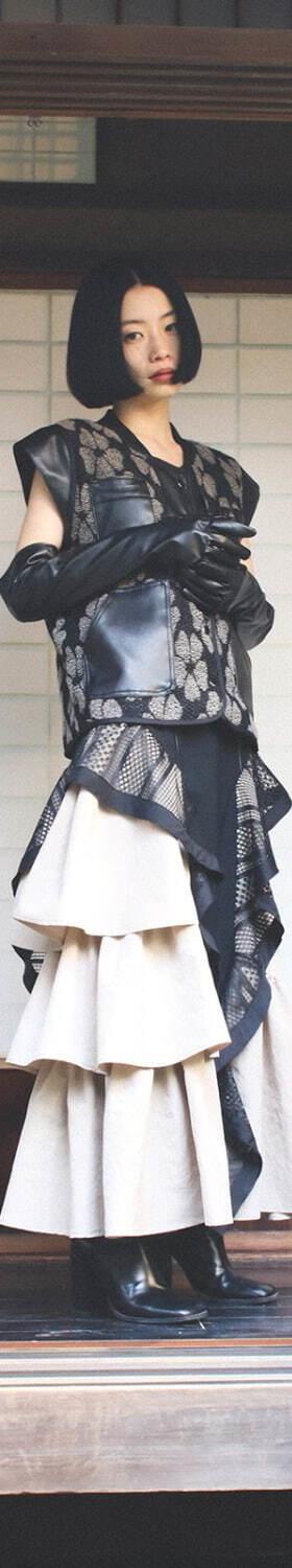 A woman wearing a sheer black lace layered vest over a tiered ivory skirt, standing on a traditional Japanese wooden veranda