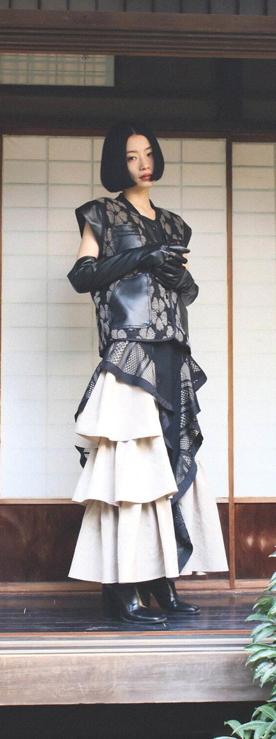 A woman wearing a sheer black lace layered vest over a tiered ivory skirt, standing on a traditional Japanese wooden veranda