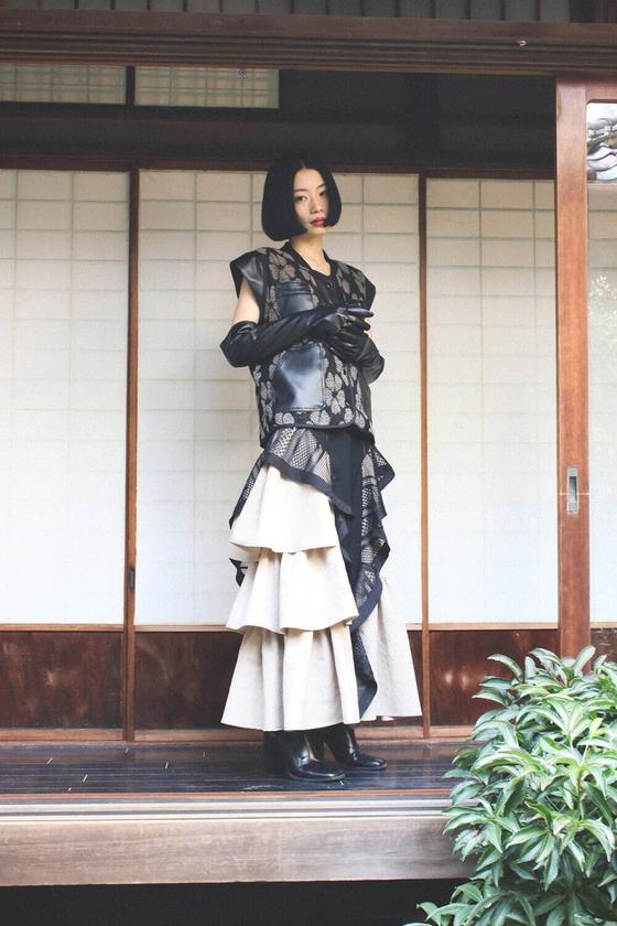 A woman wearing a sheer black lace layered vest over a tiered ivory skirt, standing on a traditional Japanese wooden veranda