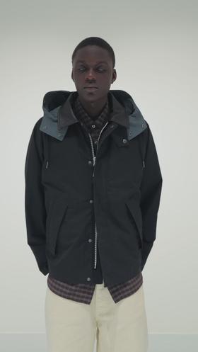 A man wearing a black Garment Project hooded jacket over a checked shirt and cream trousers