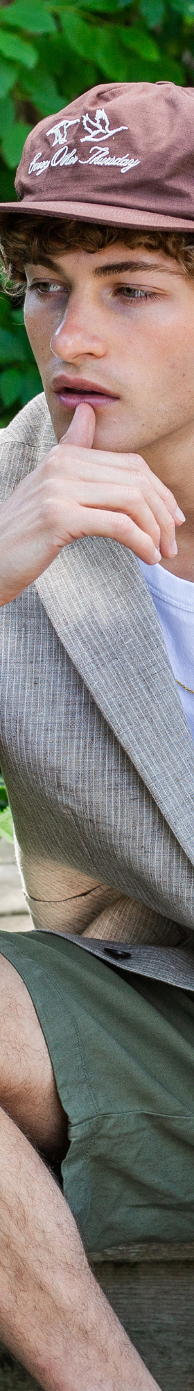 A man wearing a light grey linen blazer, white tank top and olive shorts with a brown cap, seated outdoors among ferns