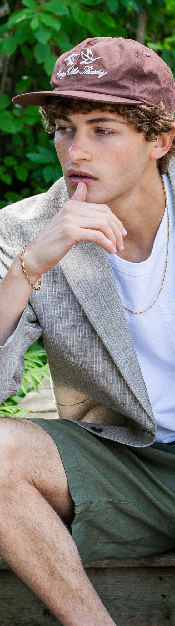 A man wearing a light grey linen blazer, white tank top and olive shorts with a brown cap, seated outdoors among ferns