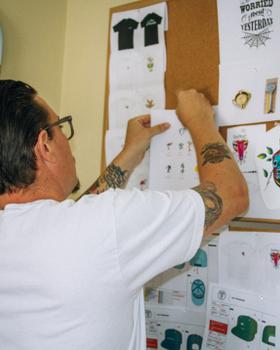 A man in a white T-shirt pinning Good Worth & Co. design sketches to a studio corkboard