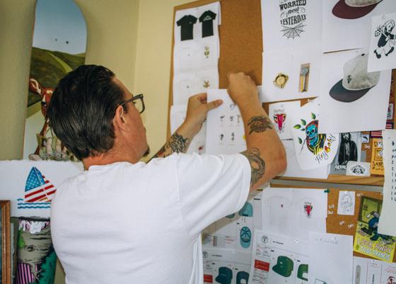 A man in a white T-shirt pinning Good Worth & Co. design sketches to a studio corkboard