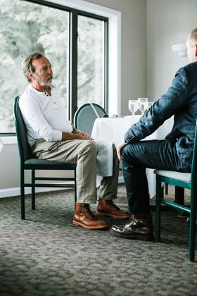 Two men seated at a table, one wearing brown leather lace-up boots with khaki trousers and a white henley