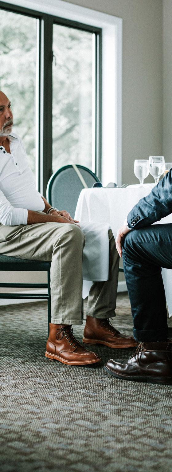Two men seated at a table, one wearing brown leather lace-up boots with khaki trousers and a white henley