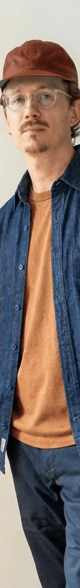 A man wearing an indigo denim work shirt over a rust tee with navy chinos