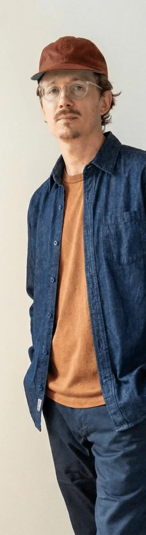 A man wearing an indigo denim work shirt over a rust tee with navy chinos