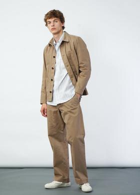 A man wearing a tan Hansen Garments chore jacket over a striped shirt with wide beige trousers