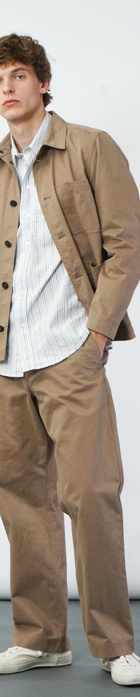 A man wearing a tan Hansen Garments chore jacket over a striped shirt with wide beige trousers