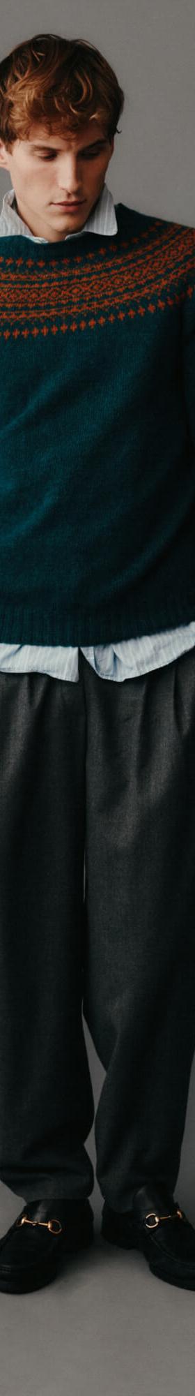 A man wearing a teal Fair Isle yoke sweater by Harley of Scotland with a blue shirt and grey pleated trousers