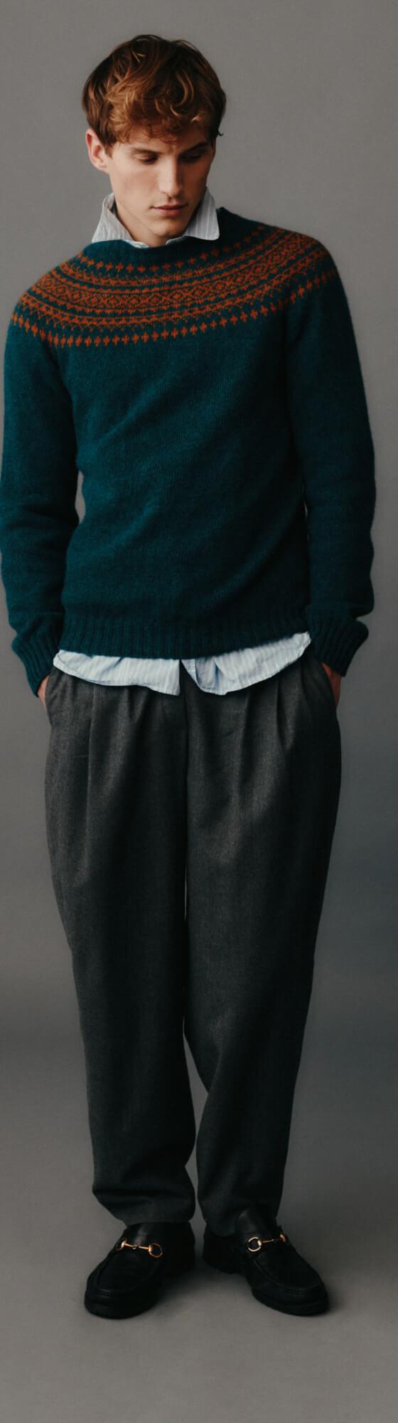 A man wearing a teal Fair Isle yoke sweater by Harley of Scotland with a blue shirt and grey pleated trousers