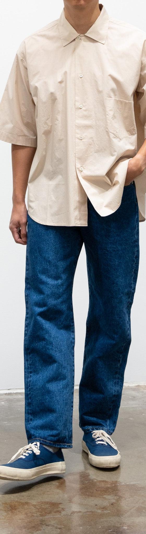 A man wearing wide-leg HATSKI selvedge jeans with an ecru short-sleeve shirt and navy sneakers