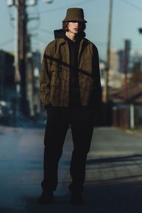 A man wearing an olive Haven field jacket, bucket hat and black trousers on a city street