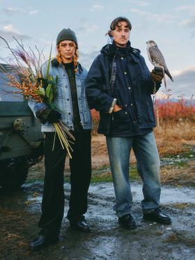 Two people standing in a field at dusk, one in a light wash denim jacket holding dried flowers, the other in a navy waxed field jacket holding a falcon