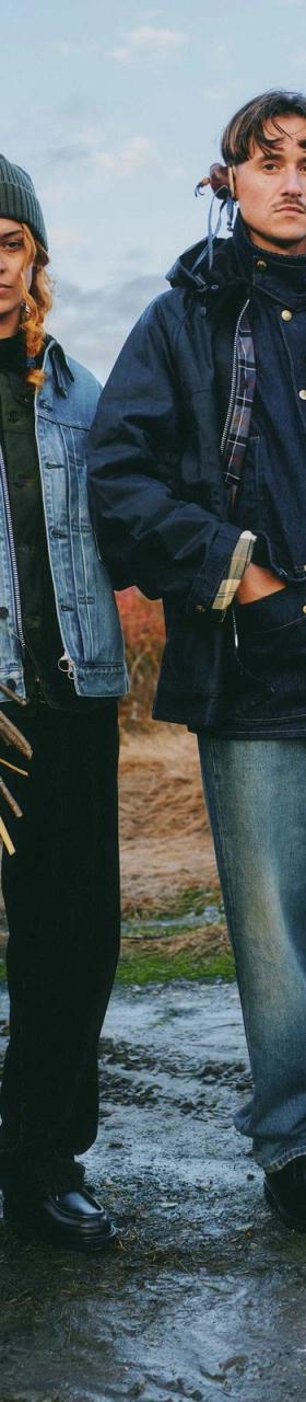 Two people standing in a field at dusk, one in a light wash denim jacket holding dried flowers, the other in a navy waxed field jacket holding a falcon