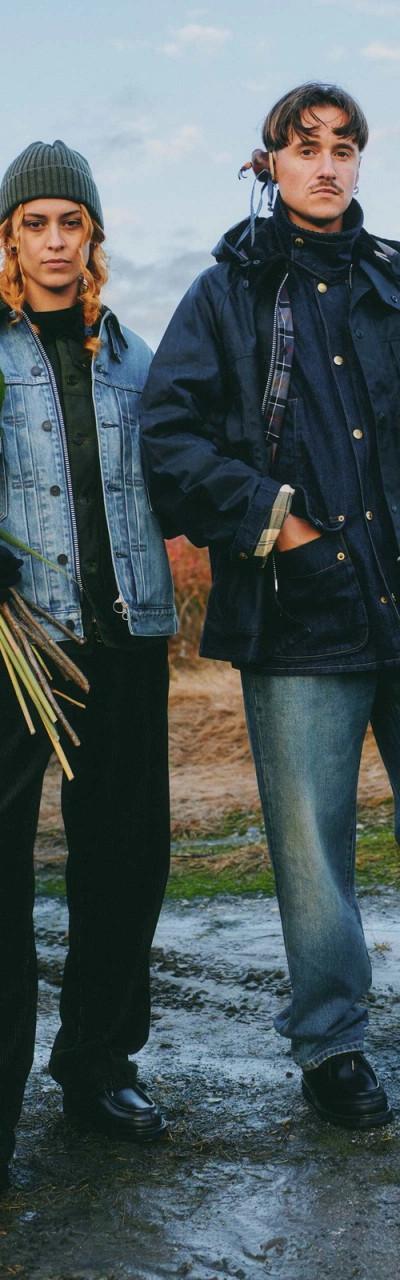 Two people standing in a field at dusk, one in a light wash denim jacket holding dried flowers, the other in a navy waxed field jacket holding a falcon