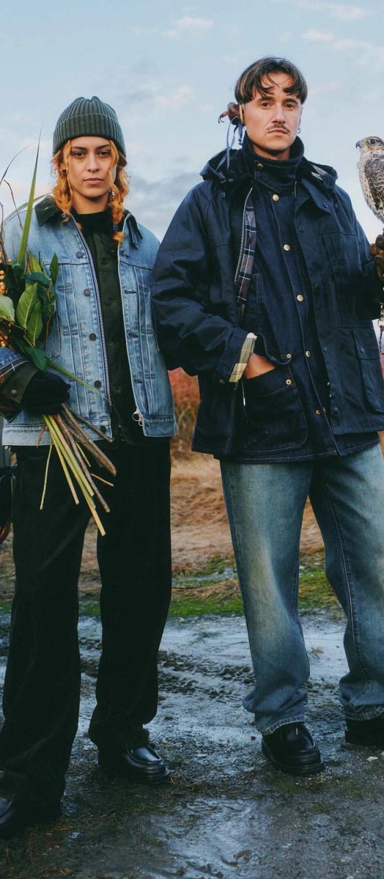 Two people standing in a field at dusk, one in a light wash denim jacket holding dried flowers, the other in a navy waxed field jacket holding a falcon