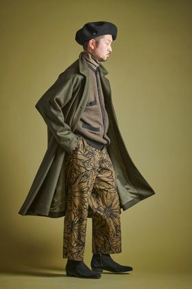 A man wearing a long olive Haversack coat over a knit jacket with floral-print wide trousers and a black beret