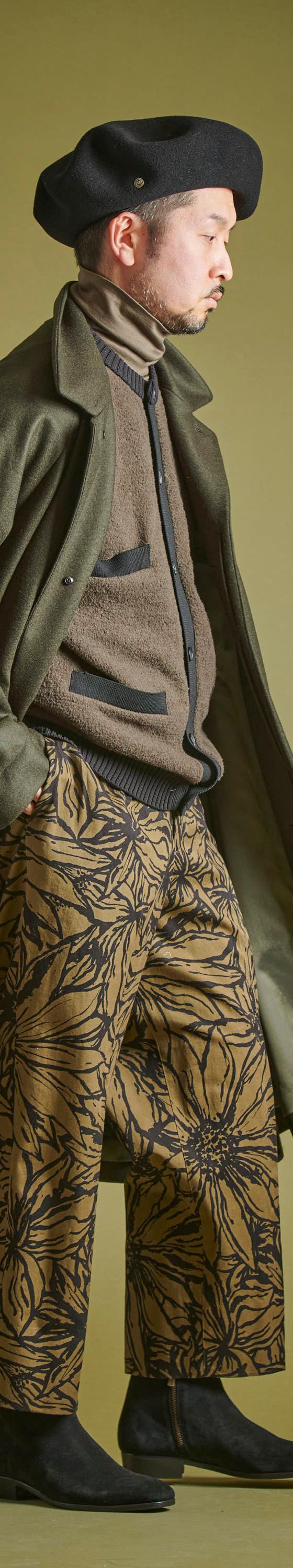 A man wearing a long olive Haversack coat over a knit jacket with floral-print wide trousers and a black beret