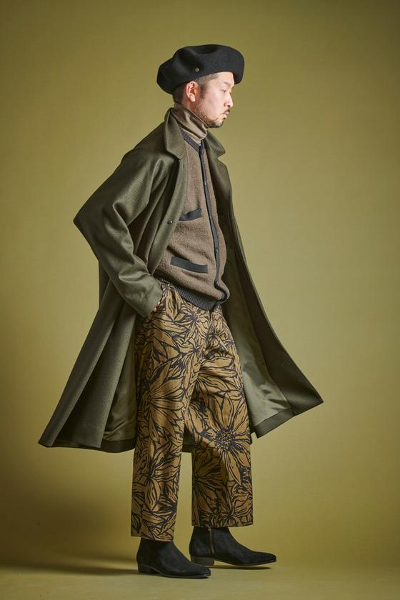 A man wearing a long olive Haversack coat over a knit jacket with floral-print wide trousers and a black beret