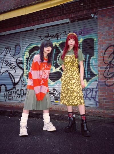 Two women wearing Heaven by Marc Jacobs looks — a red and pink striped graphic knit over a pleated skirt with platform boots, and a gold floral slip dress over an olive polo — standing on a graffiti-covered street
