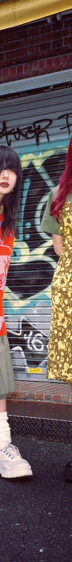 Two women wearing Heaven by Marc Jacobs looks — a red and pink striped graphic knit over a pleated skirt with platform boots, and a gold floral slip dress over an olive polo — standing on a graffiti-covered street