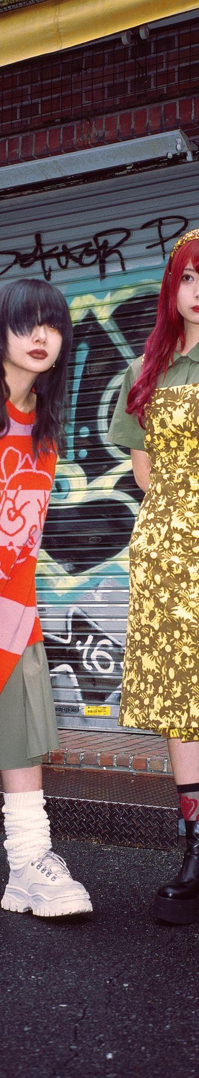 Two women wearing Heaven by Marc Jacobs looks — a red and pink striped graphic knit over a pleated skirt with platform boots, and a gold floral slip dress over an olive polo — standing on a graffiti-covered street