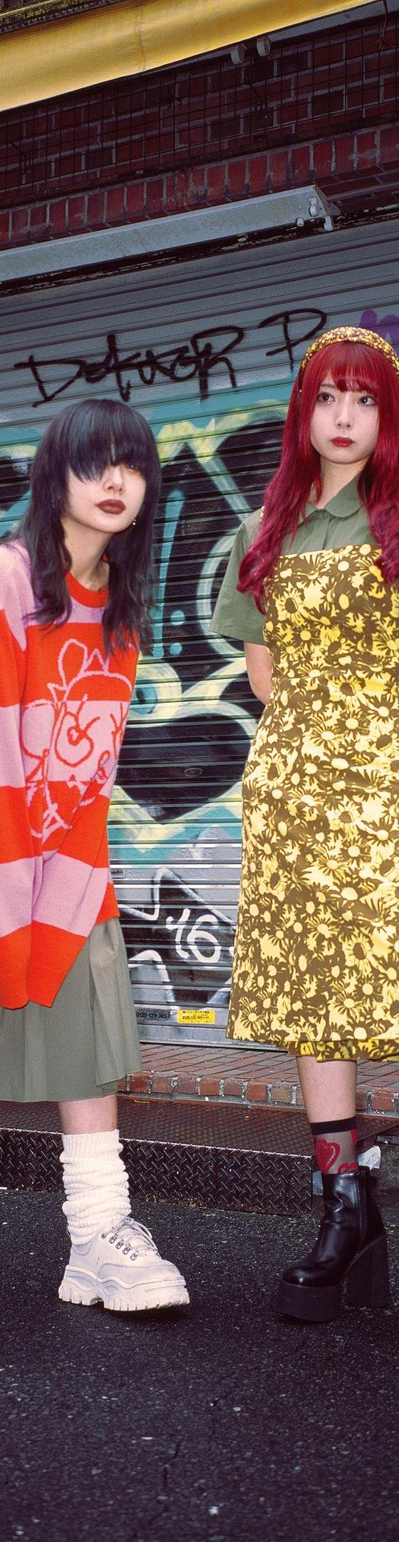 Two women wearing Heaven by Marc Jacobs looks — a red and pink striped graphic knit over a pleated skirt with platform boots, and a gold floral slip dress over an olive polo — standing on a graffiti-covered street