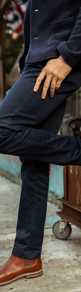 A man wearing tan leather HELM Chelsea boots with navy trousers and a navy sweater, leaning against a rusted truck