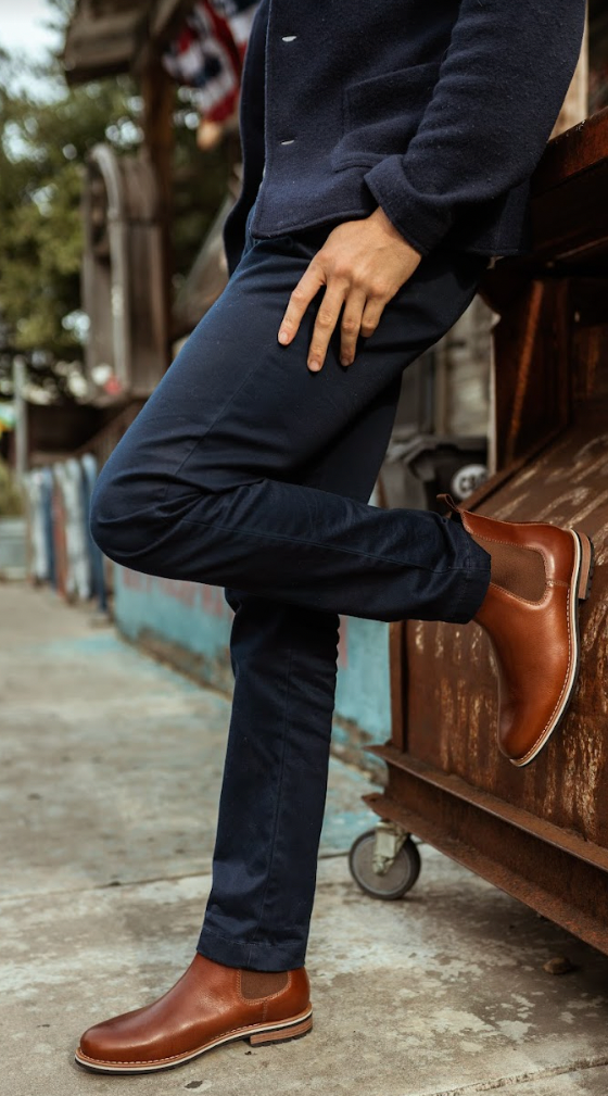 A man wearing tan leather HELM Chelsea boots with navy trousers and a navy sweater, leaning against a rusted truck