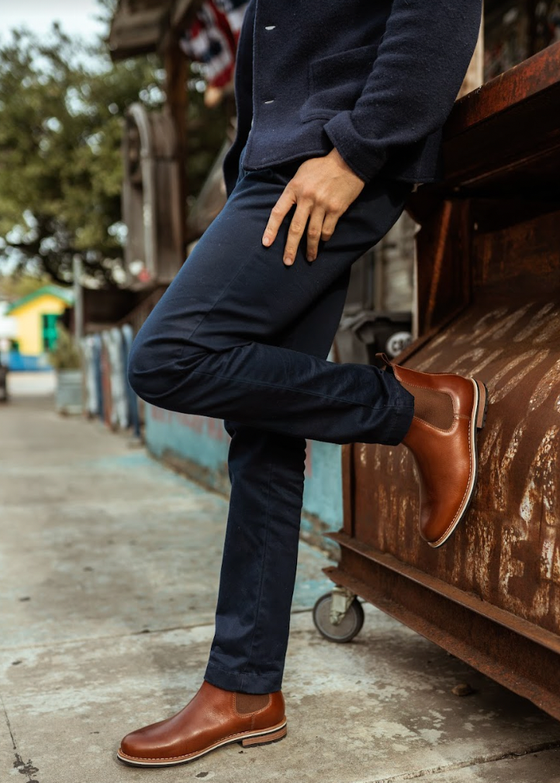 A man wearing tan leather HELM Chelsea boots with navy trousers and a navy sweater, leaning against a rusted truck