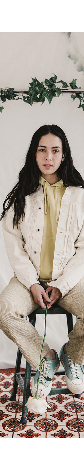 A woman seated wearing a cream corduroy jacket, olive shirt and natural leather sneakers