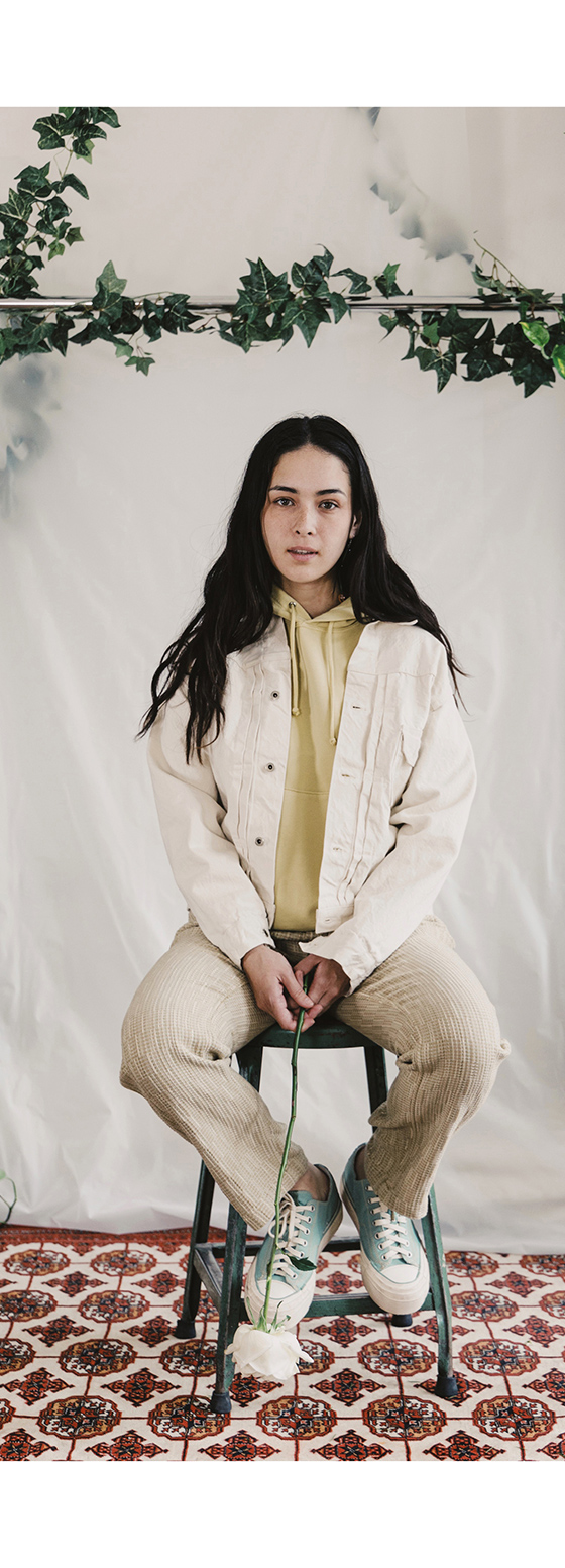 A woman seated wearing a cream corduroy jacket, olive shirt and natural leather sneakers